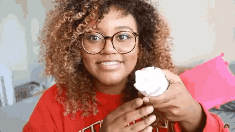 a woman with curly hair and glasses is holding a cupcake in her hand .