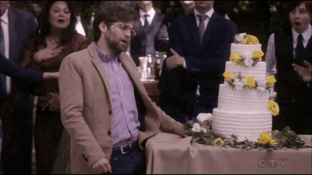 a man in a tan jacket stands in front of a wedding cake with yellow flowers