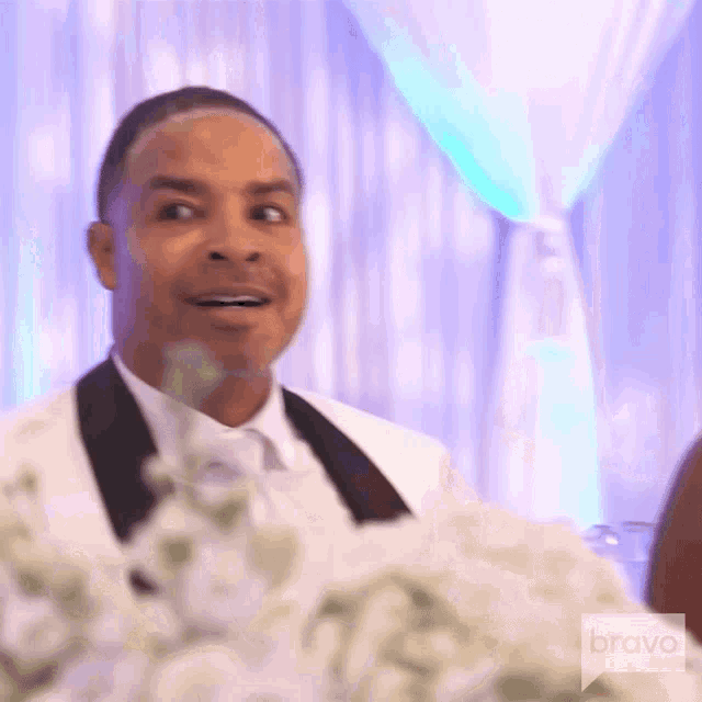 a man in a tuxedo is sitting at a table with flowers and making a funny face .