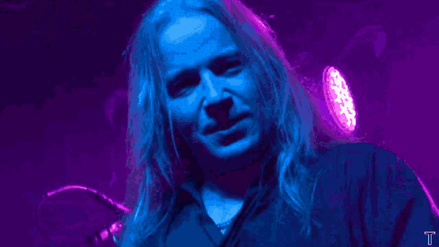 a man with long blonde hair is screaming in front of a purple light with the letter t in the lower right corner