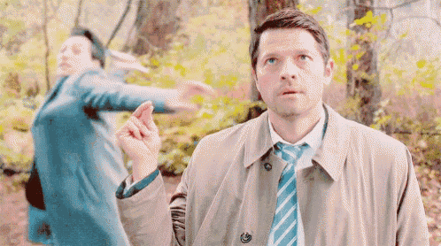 a man in a trench coat and tie is standing in the woods with a woman in a blue coat behind him .