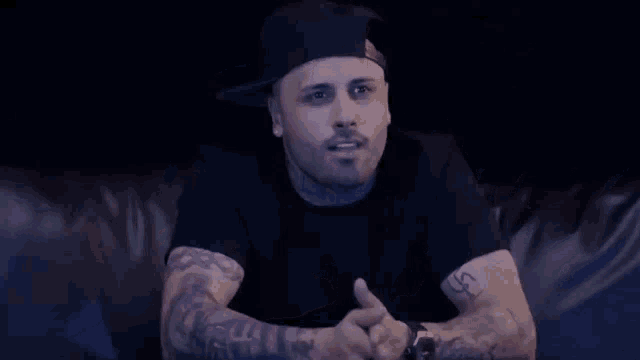 a man with a lot of tattoos is sitting on a couch with his hands folded .