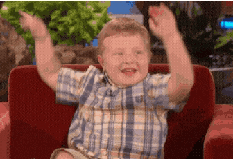 a young boy in a plaid shirt is sitting on a red couch with his arms in the air .