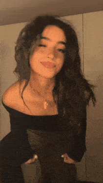 a woman in a black off the shoulder top and green pants is standing in front of a wall