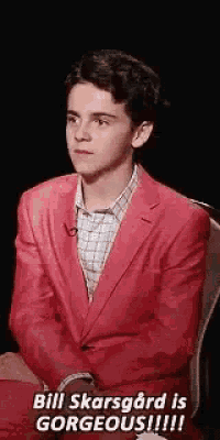 a young man in a red suit is sitting in a chair and saying `` bill skarsgard is gorgeous !!! ''