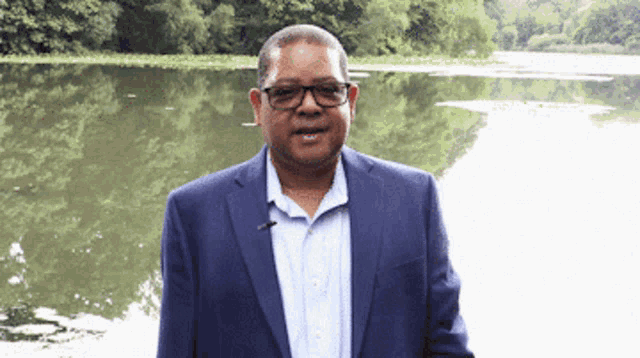 a man wearing glasses and a blue suit stands in front of a lake