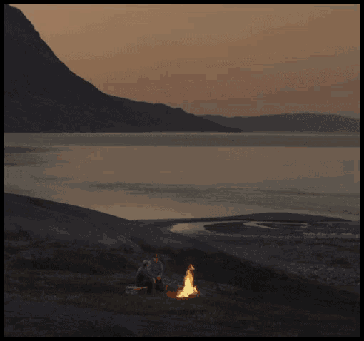 two people sit around a campfire near a lake