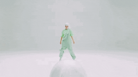 a woman in a green shirt and white wig is dancing in front of a white background .