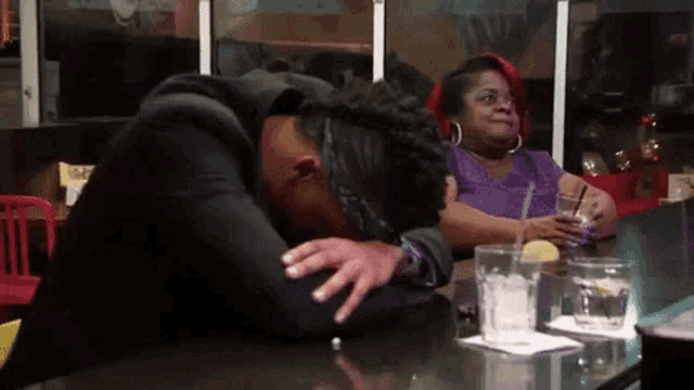 a woman is sitting at a table with a drink in her hand and a man is laying on the table with his head down .
