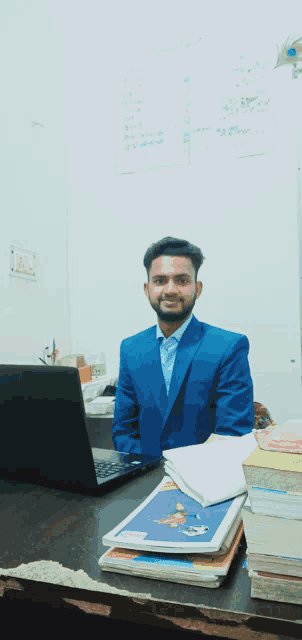 a man in a blue suit sits at a desk in front of a laptop