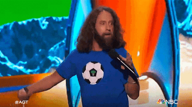 a man with a beard is holding a microphone and wearing a blue shirt with a soccer ball on it .