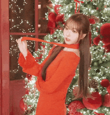 a woman in a red dress is standing in front of a christmas tree holding a red ribbon around her neck