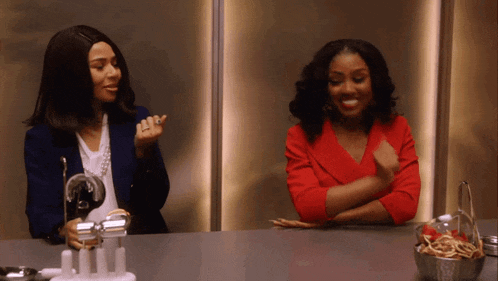 two women are sitting at a table with their arms crossed and one is wearing a red top