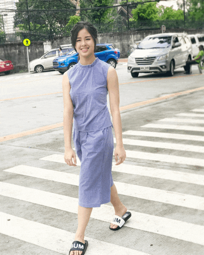 a woman wearing a purple dress and black slides with slim on them crosses a crosswalk