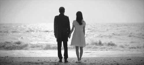 a man and a woman are standing on a beach holding hands looking at the ocean .