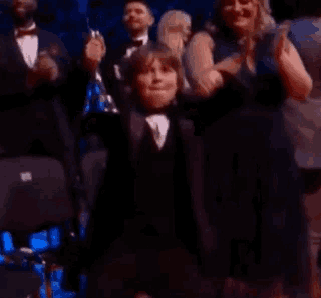 a young boy in a tuxedo is sitting in front of a crowd of people applauding .