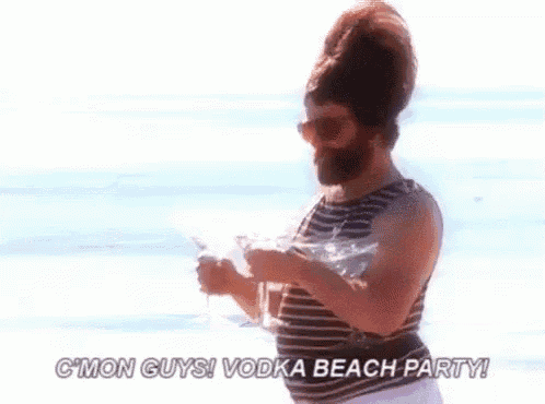 a man with a beard is standing on a beach holding a glass of vodka and a martini .