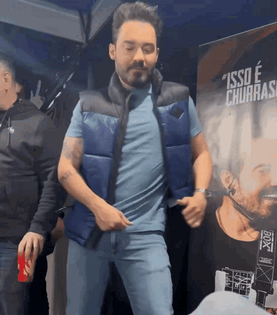 a man in a blue vest is dancing in front of a poster that says isso e churras