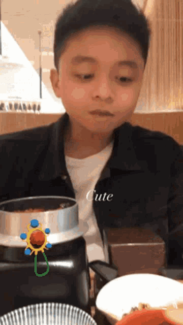 a young boy sits at a table with a bowl of food and a sticker that says cute on it