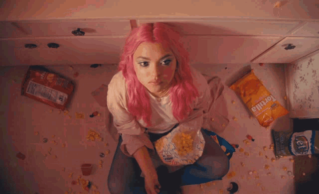 a woman with pink hair is eating tortilla chips in a messy room