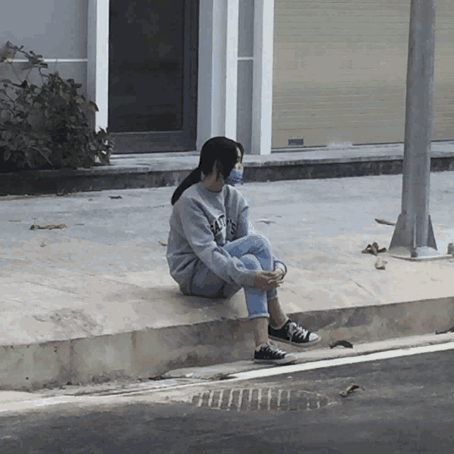 a girl wearing a mask sits on a curb wearing a sweatshirt that says ' nyc ' on the front