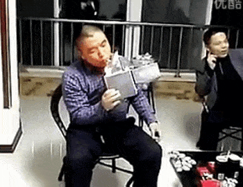 a man is sitting in a chair holding a box in his hand while another man talks on a cell phone .