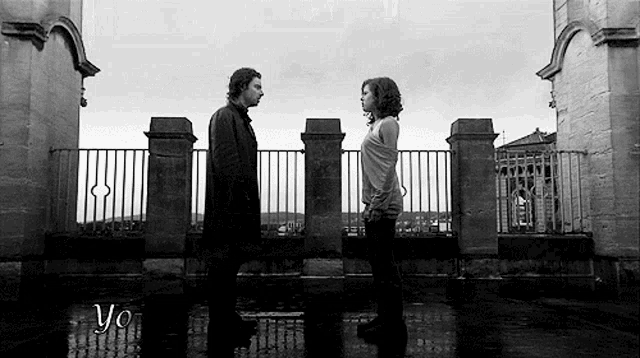 a black and white photo of a man and a woman standing next to each other in front of a fence .