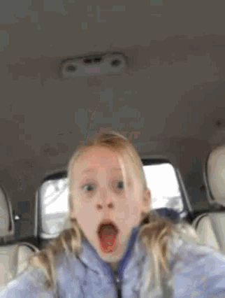 a little girl is taking a selfie in the back seat of a car with her mouth wide open .
