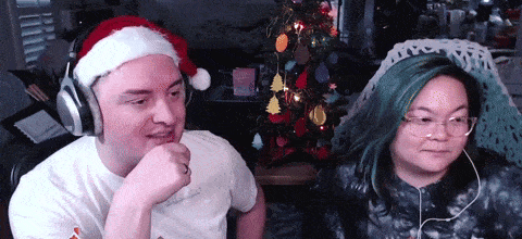 a man wearing headphones and a santa hat sits next to a woman with green hair in front of a christmas tree