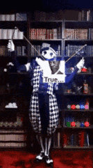 a man in a clown costume is holding a violin in front of a bookshelf that says true