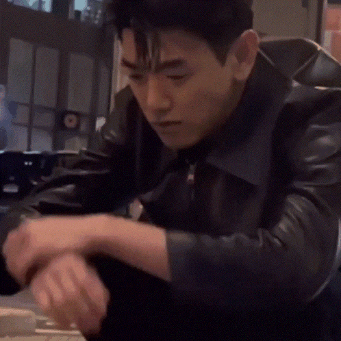 a man in a black leather jacket sits at a table with his hands folded