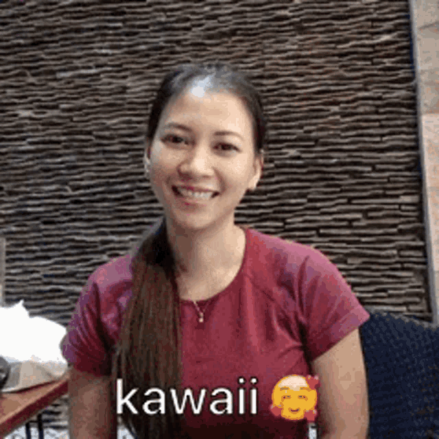 a woman in a red shirt is smiling in front of a stone wall and has the word kawaii on her shirt .