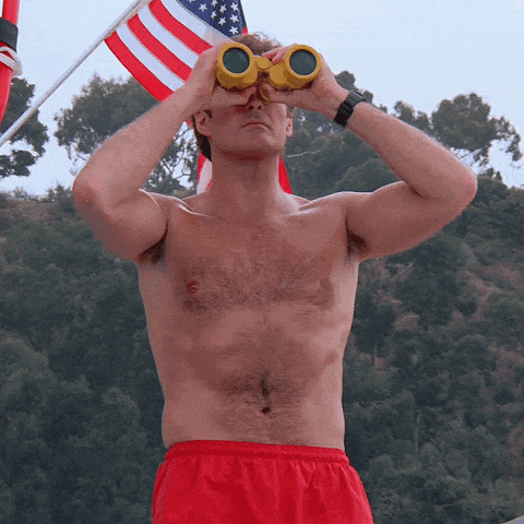 a shirtless man in red shorts looks through binoculars in front of an american flag