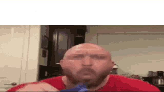 a bald man with a beard is sitting in a kitchen eating a bag of chips .