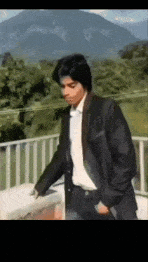 a man in a suit stands on a balcony with mountains in the background .