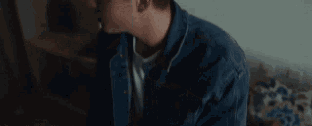 a young man wearing a denim jacket and ear buds is sitting on a bed .