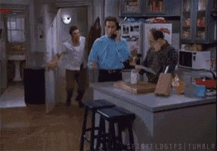 a man is talking on a cell phone while another man dances in a kitchen