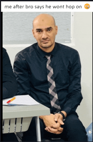 a man in a black shirt and tie sits at a desk with his hands folded
