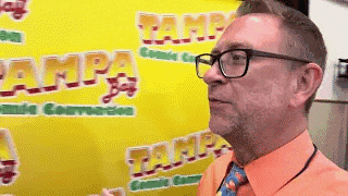 a man wearing glasses and an orange shirt is standing in front of a yellow banner that says tampa comic convention .