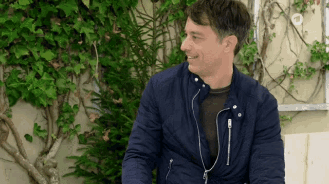 a man in a blue jacket is smiling in front of a wall with ivy