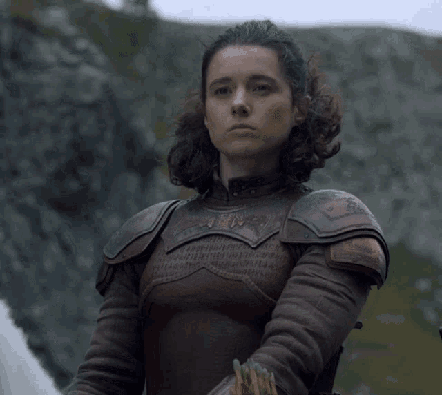 a woman in armor stands in front of a mountain and looks at the camera