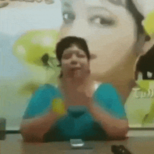 a woman in a blue shirt is sitting at a table in front of a picture of a woman 's face .