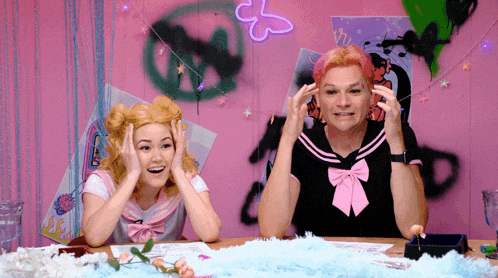 a man and a woman are sitting at a table with a pink wall behind them