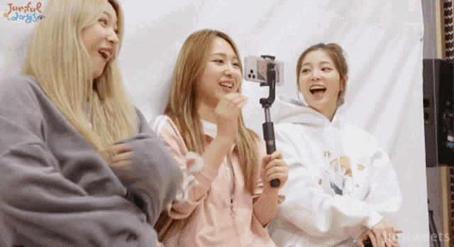 three girls are laughing and one is holding a cell phone in front of a wall that says joyful days