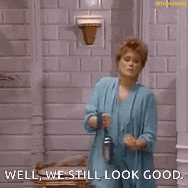 a woman in a blue robe is standing in front of a brick wall and says `` well , we still look good '' .