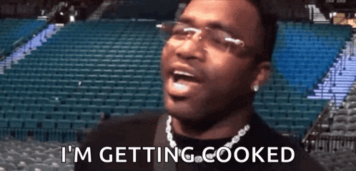 a man wearing glasses and a necklace is saying i 'm getting cooked in front of a stadium .