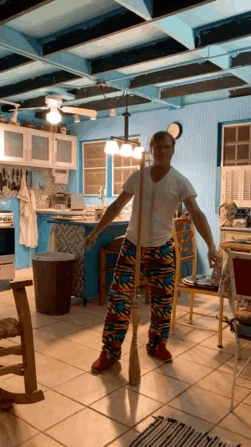 a man in a white shirt and colorful pants is standing in a kitchen holding a mop