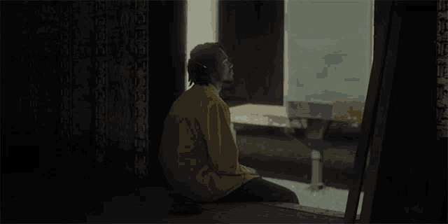a man in a yellow jacket sits on the floor in front of a window