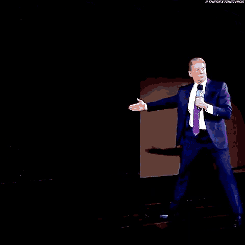 a man in a suit and tie is standing on a stage holding a microphone while another man watches