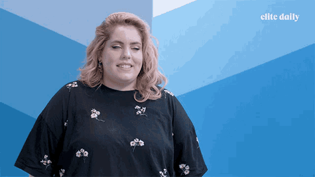 a woman in a black shirt is smiling in front of a blue wall with elite daily written on it